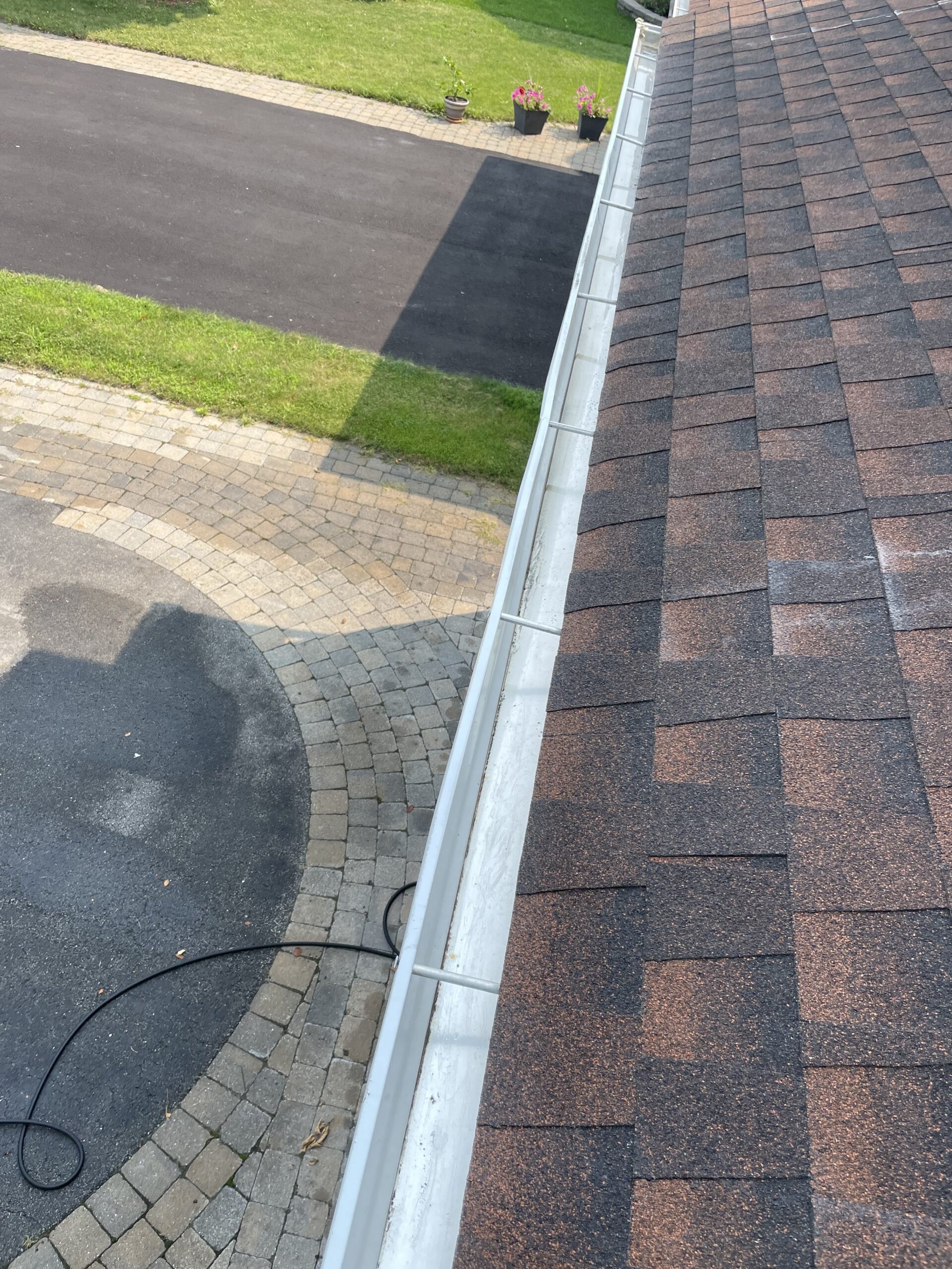 Overhead view along a clean white rain gutter on a shingled roof, looking down toward a driveway and landscaped front yard